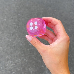 May include: A pink, translucent dice with white dots. The dice is being held in a hand.