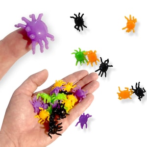 Op de afbeelding: Een verzameling kleine, plastic, spinvormige speelgoedjes in verschillende kleuren, waaronder paars, zwart, groen en oranje. De spinnen zijn verspreid over een wit oppervlak en in een hand.