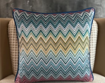 Puede incluir: Cojín decorativo cuadrado con un colorido patrón de zigzag en tonos turquesa, amarillo, rosa y blanco. El cojín tiene un borde azul oscuro y está colocado sobre una silla beige.