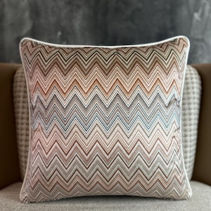 Może przedstawiać: Kwadratowa poduszka dekoracyjna z zygzakowatym wzorem w odcieniach brązu, beżu i niebieskiego. Ma białą obwódkę i jest umieszczona na neutralnej powierzchni. Element dekoracji wnętrz.