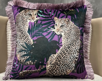 Op de afbeelding: Vierkant decoratief kussen met een luipaardprint. Het kussen heeft een paarse achtergrond met groene bladeren en twee luipaarden. De randen zijn afgewerkt met een paarse franje. Het kussen ligt op een grijze ondergrond.