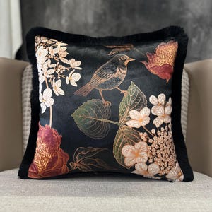May include: Black decorative pillow with a bird and floral design. The pillow features a detailed illustration of a bird perched on a branch, surrounded by flowers and leaves in shades of white, peach, and gold. The pillow has a black fringe border.