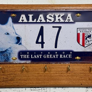May include: A wooden key holder with a metal plaque featuring a blue and white Alaskan license plate with the text "ALASKA 47 IDITAROD THE LAST GREAT RACE" and a white husky dog.