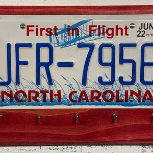 May include: A red wooden key holder with a North Carolina license plate featuring the text "First in Flight" and the license plate number "JFR-7956".