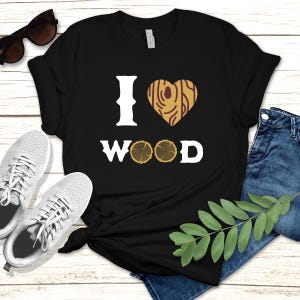 以下が含まれることがあります： 「I ♥ WOOD」の文字が入った黒いTシャツ。ハートのグラフィックは木目模様です。「WOOD」の文字はハートと「I」の下にあります。このシャツは、グレーのスニーカーとブルージーンズと組み合わせています。