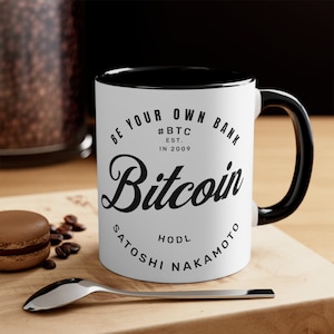 May include: White ceramic mug with a black rim and handle. The mug has a black graphic that says "Be Your Own Bank #BTC Est. In 2009 Bitcoin HODL Satoshi Nakamoto".