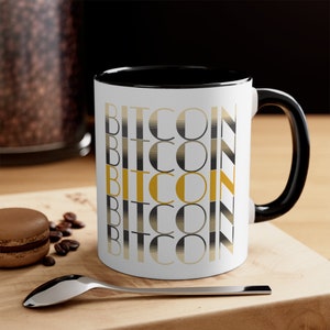 May include: White ceramic mug with a black rim and handle. The mug has a design of the word "Bitcoin" repeated vertically in a black and gold striped pattern.