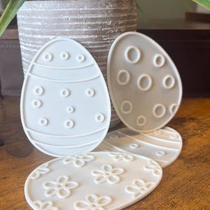 May include: White, egg-shaped coasters with various designs. Some feature dotted patterns, others have stripes, and one has a floral design. These decorative coasters are perfect for Easter or spring decor.