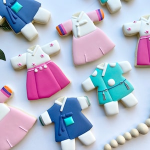 May include: Seven decorated sugar cookies shaped like traditional Korean hanbok clothing. The cookies are in various colors, including blue, pink, white, and green.