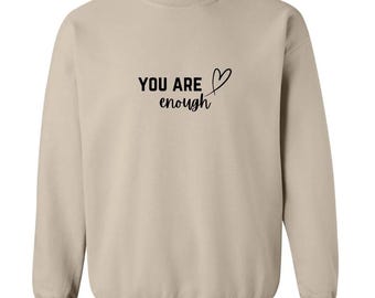 Op de afbeelding: Een beige sweatshirt met de tekst "YOU ARE enough" en een kleine hartgrafiek gedrukt op de voorkant.