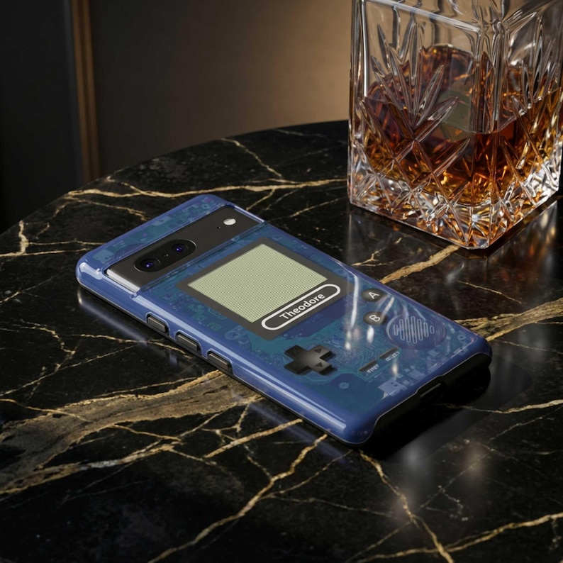 A blue phone case with a retro video game design, featuring a screen, buttons, and a custom name. The case is on a black marble surface, with a decanter in the background. iPhone 17 Pro Max case, Samsung s26 Ultra case, Pixel 9 Pro case, and more.
