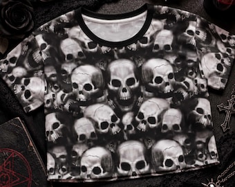 May include: A short-sleeved crew neck t-shirt featuring an all-over repeating graphic print of realistic human skulls in shades of white, gray, and black. The garment has a solid black ribbed neckline and a straight hemline, showcasing a detailed pattern.