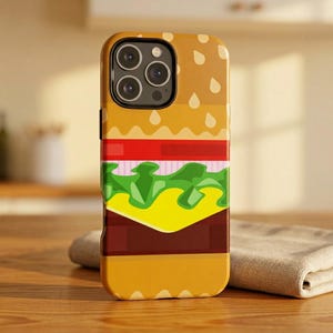 May include: A smartphone case featuring a graphic design of a cheeseburger. The design includes layers of a sesame seed bun, red tomato, light pink onion, green lettuce, yellow cheese, and a brown burger patty, all in a flat, stylized illustration.