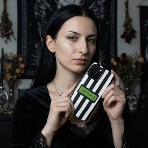 An gothic woman holding a custom black and white striped phone case with the name 'CATHERINE'. The custom phone case is available for iPhone, Samsung Galaxy and Google Pixel and is a perfect personalized gift for her.