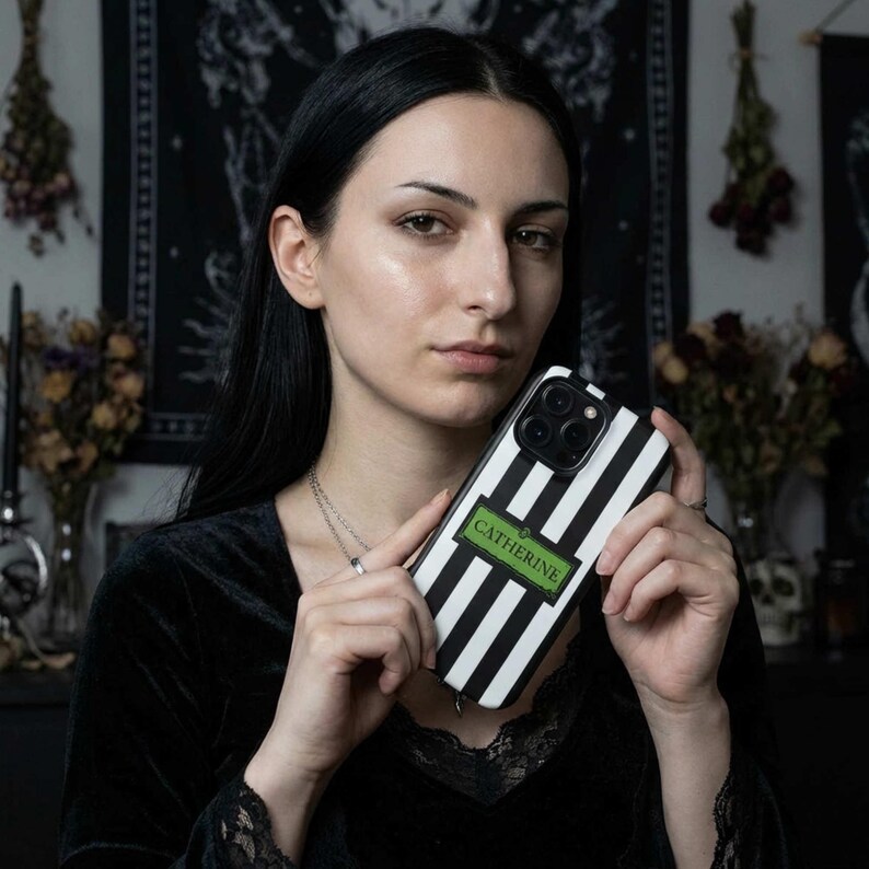 An gothic woman holding a custom black and white striped phone case with the name 'CATHERINE'. The custom phone case is available for iPhone, Samsung Galaxy and Google Pixel and is a perfect personalized gift for her.