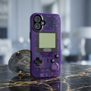 May include: A transparent purple phone case with a retro video game design. The case features a screen, buttons, and the text "Pretty Rebel". The case is on a black and gold marble surface.