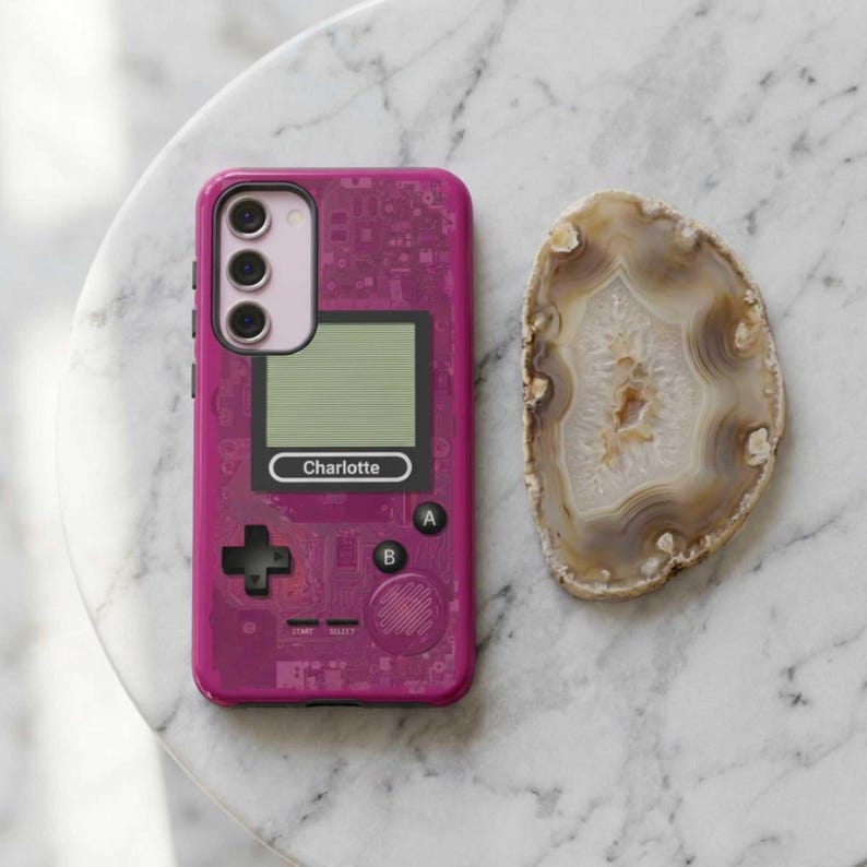 A pink phone case with a retro video game design, featuring a screen, buttons, and a custom name. The phone cover is on a white and gray marble surface and next to it is a polished, oval-shaped agate stone with brown and cream-colored bands. Phone case for iPhone, Samsung and Pixel. Personalized gift for him, gamers and Nintendo fans.