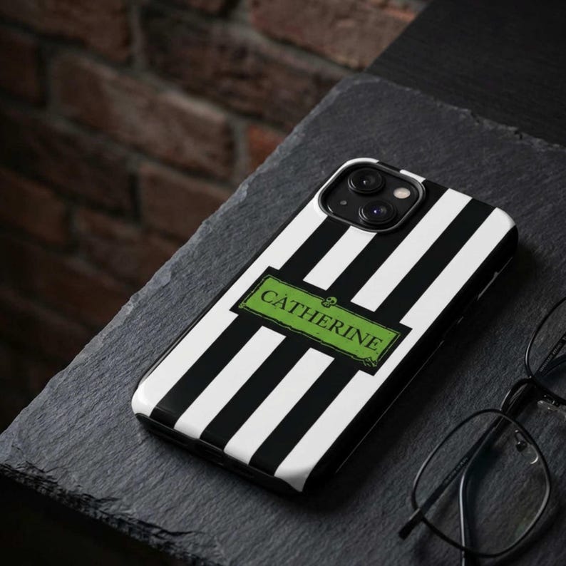 A custom black and white striped phone case with the name "Catherine" in a green rectangle. The case is on a dark gray surface, with a brick wall in the background. The custom phone case is available for iPhone, Samsung Galaxy and Google Pixel and is a perfect personalized gift for her.