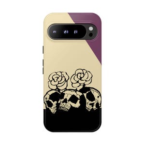 May include: A phone case with a unique design featuring three skulls with floral accents. The case has a color block design with beige, purple, and black sections. The phone's camera is visible at the top.
