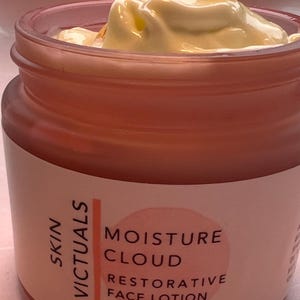 Puede incluir: Un tarro rosa de crema facial Skin Victuals Moisture Cloud Restorative Face Lotion con una cuchara de madera que saca la crema. El tarro está etiquetado con "Skin Victuals Moisture Cloud Restorative Face Lotion" y "Net Wt 2 oz."
