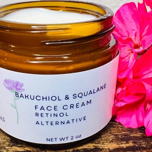 Puede incluir: Un tarro de vidrio marrón con una etiqueta blanca que dice "Bakuchiol & Squalane Crema Facial Alternativa al Retinol" y "Peso Neto 2 oz". El tarro está lleno de una crema blanca y está sobre una superficie de madera.
