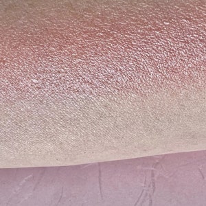 Pu&ograve; includere: Primo piano di un ombretto iridescente scintillante con un gradiente rosa e bianco. L'ombretto ha una texture fine e glitterata.