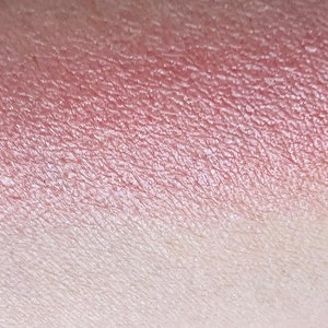 Pu&ograve; includere: Un primo piano di un campioncino di ombretto rosa e bianco sulla pelle. L'ombretto ha una finitura scintillante.