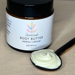 May include: A white jar with a black lid labeled "Skinvictuals Body Butter Neroli Cream" with a net weight of 4 oz. A small spoonful of white body butter is on a black and white wooden spoon.