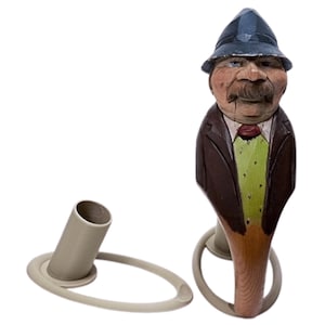 May include: A whimsical, hand-carved wooden bottle stopper featuring a man's face with a mustache and hat. The stopper has a brown suit, green shirt, and red tie. A separate, light gray bottle holder is also included.