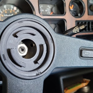 Puede incluir: Primer plano de un volante negro con un cubo central circular de varias capas. El volante está en el interior de un coche con un salpicadero y medidores visibles en el fondo.