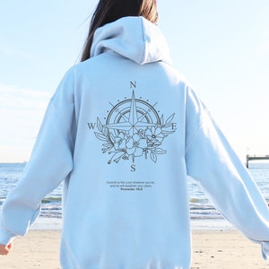 Op de afbeelding: Een lichtblauwe hoodie met een kompasontwerp en bloemmotieven op de rug. Het kompas wijst naar het noorden, zuiden, oosten en westen. De tekst "Vertrouw de Heer in al wat je doet, en hij zal je plannen laten slagen." Spreuken 16:3 staat onder het kompas.