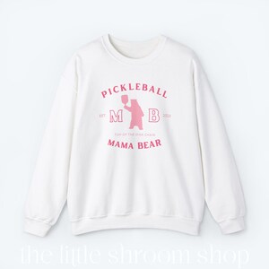 Op de afbeelding: Wit sweatshirt met een roze grafisch ontwerp met de tekst "Pickleball" en "Mama Bear". De afbeelding bevat ook een roze beer die een pickleball-racket vasthoudt.