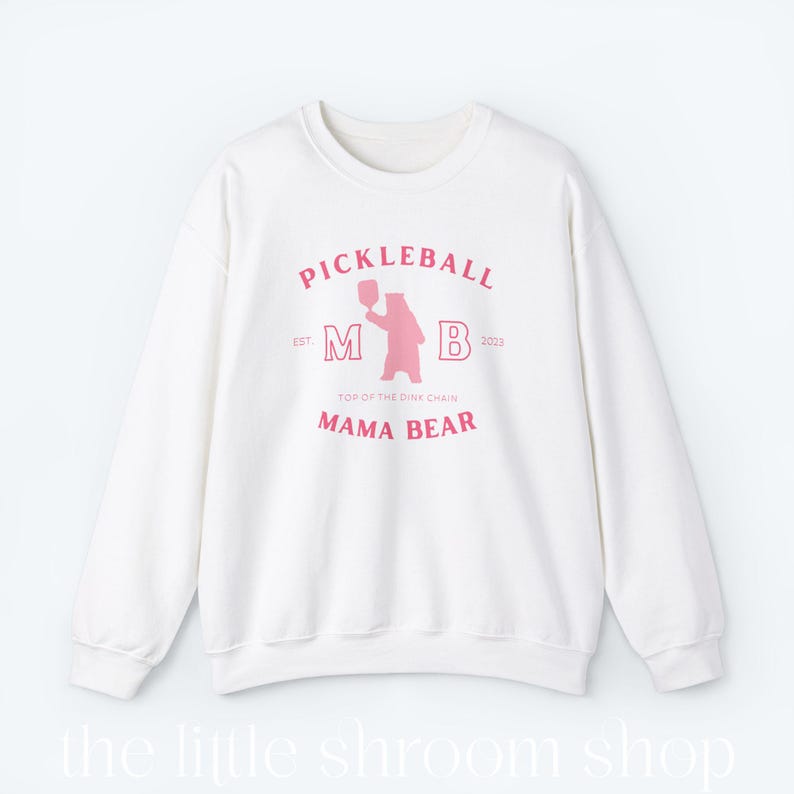 Op de afbeelding: Wit sweatshirt met een roze grafisch ontwerp met de tekst "Pickleball" en "Mama Bear". De afbeelding bevat ook een roze beer die een pickleball-racket vasthoudt.