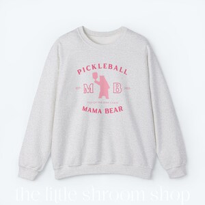 Op de afbeelding: Een grijs sweatshirt met een roze grafisch ontwerp. De grafiek toont een beer die een pickleball-racket vasthoudt met de tekst "Pickleball" boven de beer en "EST. M B 2023" onder de beer. De tekst "TOP OF THE DINK CHAIN" staat boven de tekst "MAMA BEAR".