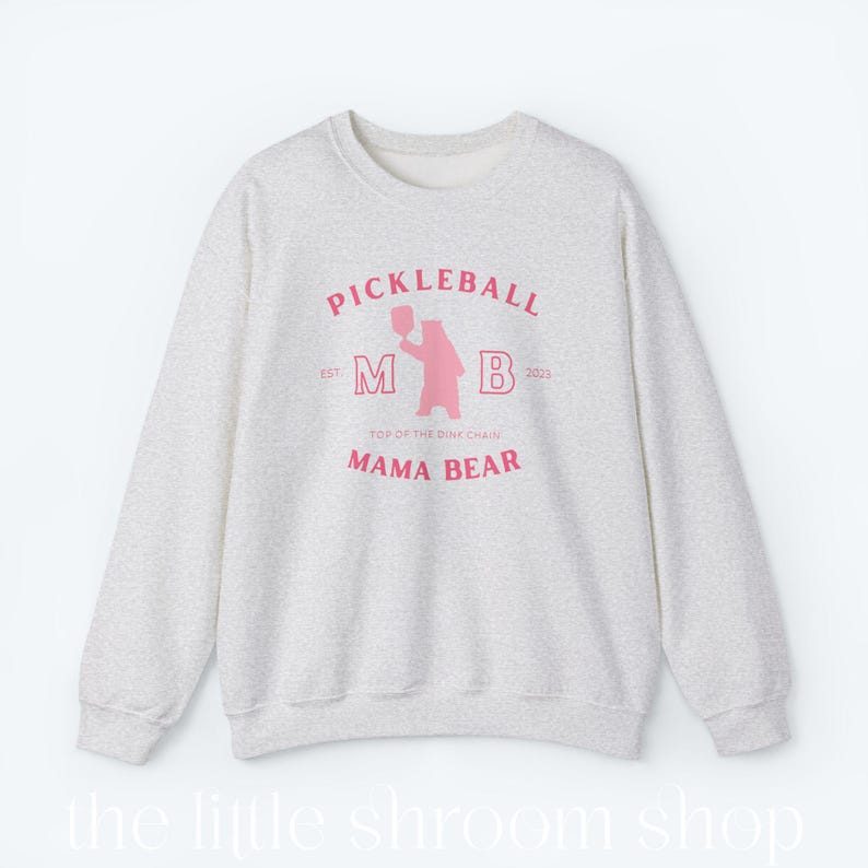 Op de afbeelding: Een grijs sweatshirt met een roze grafisch ontwerp. De grafiek toont een beer die een pickleball-racket vasthoudt met de tekst "Pickleball" boven de beer en "EST. M B 2023" onder de beer. De tekst "TOP OF THE DINK CHAIN" staat boven de tekst "MAMA BEAR".