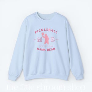 Op de afbeelding: Lichtblauwe trui met een roze afbeelding van een beer die een pickleball-racket vasthoudt. De afbeelding bevat de tekst "Pickleball", "M B", "Est. 2023", "Top of the Dink Chain" en "Mama Bear".