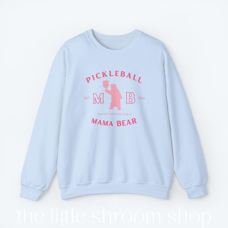 Op de afbeelding: Lichtblauwe trui met een roze afbeelding van een beer die een pickleball-racket vasthoudt. De afbeelding bevat de tekst "Pickleball", "M B", "Est. 2023", "Top of the Dink Chain" en "Mama Bear".