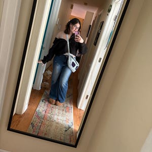 May include: A full-length mirror with a black frame reflects a person wearing a black jacket, blue jeans, and a crocheted bag with purple accents. The hallway has a colorful rug and light-colored walls.