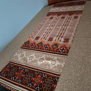 May include: A long, rectangular table runner with a geometric pattern in shades of red, orange, black, white, and green. The pattern is reminiscent of traditional folk art.