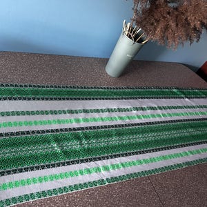 Peut inclure: Chemin de table rectangulaire avec un fond blanc et des motifs brodés verts et noirs. Le chemin de table est orné de rayures horizontales et de motifs floraux. Un vase en métal vert avec des plantes séchées est sur la table.