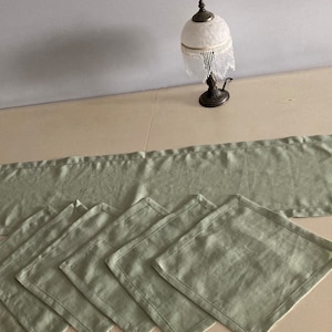 May include: A set of five sage green linen placemats and a matching table runner. The placemats and runner have a subtle texture and a simple, elegant design.