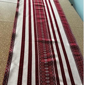 May include: White table runner with red and black stripes and a red embroidered pattern. The pattern features a repeating geometric design with small white flowers.