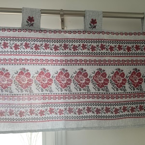 Op de afbeelding: Een witte valance met een rood en zwart geborduurd bloemenpatroon. De valance heeft een roede-opening en is gemaakt van een lichtgewicht stof.