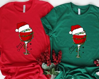 May include: Two t-shirts with a Christmas-themed design. The red shirt has a glass of wine with a Santa hat and Christmas lights on it. The green shirt has the same design but is on a green shirt. Both shirts say "Merry Christmas".