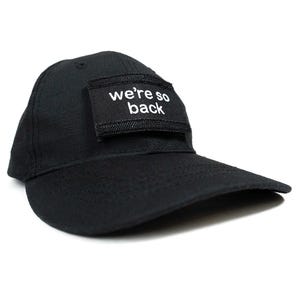 Può includere: Un cappello da baseball nero con una toppa nera che dice "we're so back" in lettere bianche.