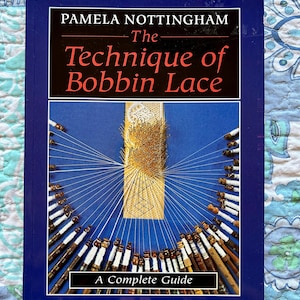 May include: A hardcover book titled "The Technique of Bobbin Lace" by Pamela Nottingham. The cover is dark blue with white and red text. The image on the cover shows a detailed view of bobbin lace with bobbins and pins. The book is a complete guide.