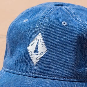 Op de afbeelding: Een blauwe denim baseball cap met een gebogen klep. De pet heeft een wit ruitvormig embleem met de tekst "PACIFIC CREST TRAIL SYSTEM" en een zeilbootafbeelding. Gemaakt van duurzaam denim materiaal.