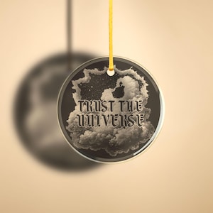 Könnte beinhalten: Ein runder Glasanhänger mit einem Schwarzweißbild von Wolken und Sternen. Der Anhänger hat eine goldene Schnur zum Aufhängen und den Text "TRUST THE UNIVERSE" in gotischer Schrift.