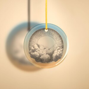 May include: A round glass ornament with a vintage illustration of a sun shining through clouds. The ornament is suspended by a yellow string.