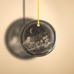 Op de afbeelding: Een ronde glazen ornament met een zwart-witte afbeelding van wolken, sterren en een halve maan. De tekst "cosmic magic" is geschreven in een gestileerd lettertype.